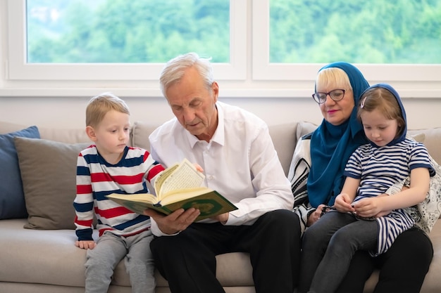 współcześni muzułmańscy dziadkowie z wnukami czytają Koran i modlą się razem na kanapie przed kolacją iftar podczas uczty ramadanu w domu