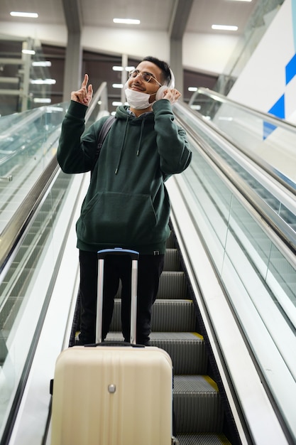 Współczesny szczęśliwy młody człowiek w casualwear, cieszący się ulubioną muzyką w słuchawkach, stojąc na stopniu ruchomych schodów ruchomych na lotnisku