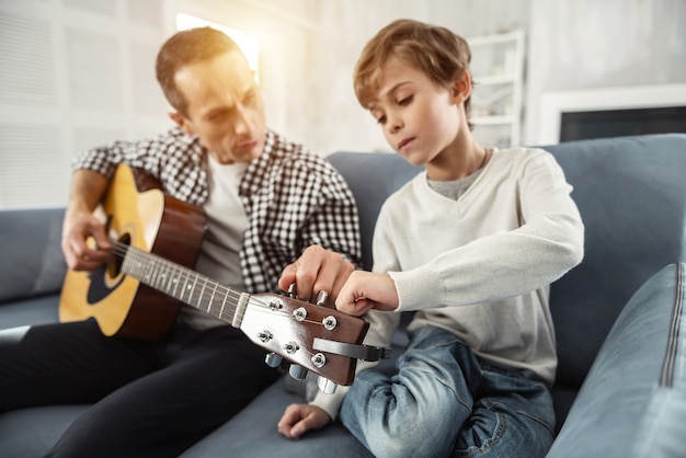 Wspólne granie. Atrakcyjny radosny dobrze zbudowany mężczyzna trzymający gitarę i siedząc na kanapie uczy swojego syna gry na gitarze