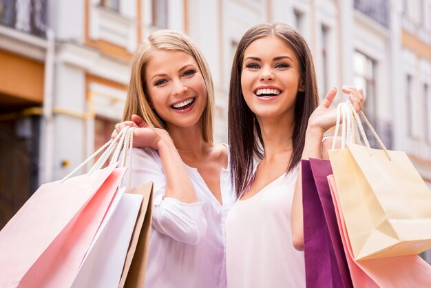 Wspólne zakupy to świetna zabawa. Dwie atrakcyjne młode kobiety trzymające torby na zakupy i uśmiechnięte stojąc na zewnątrz