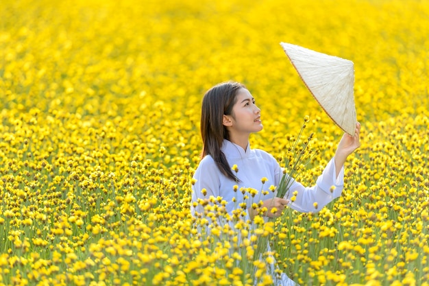 Wśród żółtych kwiatów stała kobieta w starożytnym wietnamskim stroju narodowym, trzymająca kapelusz wykonany z wikliny