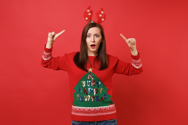 Zdjęcie wstrząśnięty, oszołomiony, młoda dziewczyna santa, wskazując palcami wskazującymi na zabawne ozdobne rogi jelenia na głowie na białym tle na czerwonym tle. szczęśliwego nowego roku 2019 celebracja party wakacje koncepcja. makieta miejsca na kopię.
