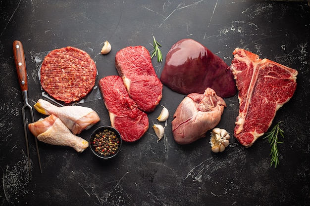 Wybór surowego mięsa z przyprawami do diety mięsożerców bez węglowodanów: niegotowany stek wołowy, kotlet z mielonego mięsa, serca, wątróbka i udka z kurczaka na czarnym kamiennym tle z góry