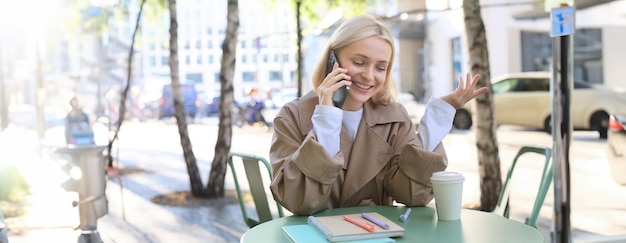 Zdjęcie wychodząca młoda szczęśliwa kobieta siedząca w kawiarni na świeżym powietrzu rozmawiająca przez telefon komórkowy uśmiechając się spędzając czas