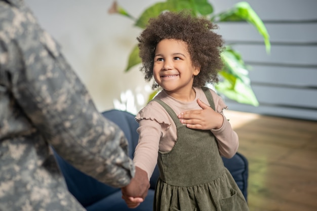 Wychowanie. Uśmiechnięta kręcona urocza afroamerykańska mała dziewczynka drży tata w mundurze kamuflażowym stojąc w domu