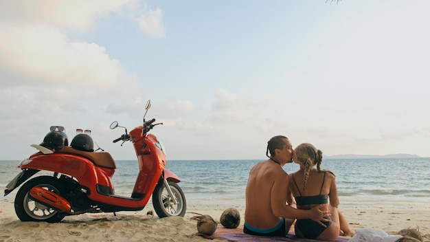 Wycieczka skuterem. Urocza para na czerwonym motocyklu w białych ubraniach na piaszczystej plaży. Świeżo poślubieni ludzie całują się uściskami spacerując w pobliżu tropikalnych palm, morza. Ślubna podróż poślubna przez ocean. Wynajem motocykli.