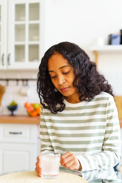 Zdjęcie wyczerpana afroamerykanka czekająca, aż pigułka musująca rozpuści się w szklance wody w kuchni