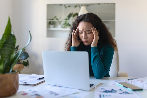 Wyczerpana azjatycka bizneswoman ma ból głowy w biurze Kreatywna kobieta pracująca przy biurku czuje się zmęczona Zestresowana przypadkowa kobieta biznesu odczuwa ból głowy podczas przepracowania na laptopie