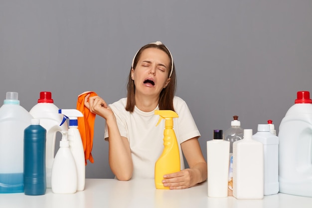 Wyczerpana kobieta pozująca w miejscu pracy z detergentami czyszczącymi odizolowanymi na szarym tle, płacząca z rozpaczy, będąc zmęczona ścinaniem zmęczenia