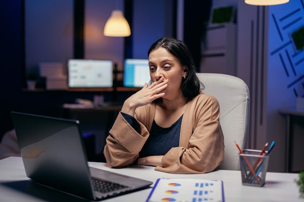 Wyczerpana kobieta ziewa pracująca nad ważnym projektem w firmie finansowej. Inteligentna kobieta siedzi w swoim miejscu pracy w późnych godzinach nocnych wykonując swoją pracę.