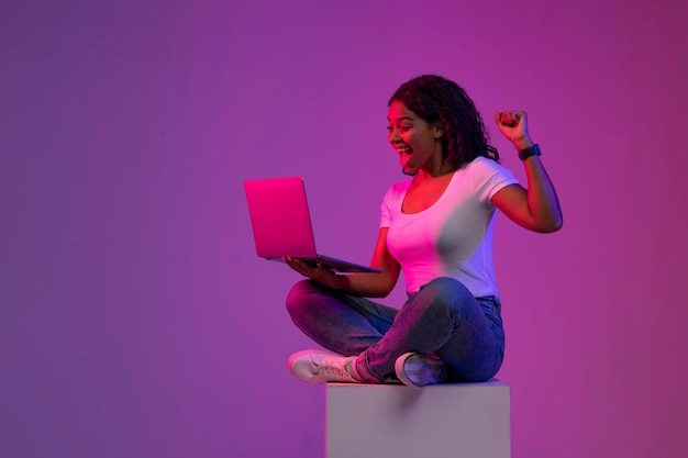 Wygrana online uszczęśliwiona czarna kobieta świętuje sukces z laptopem w świetle neonowym