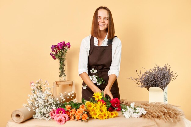 Wykwalifikowany projektant kwiatów Różne kompozycje kwiatowe Delikatne dekoracje kwiatowe Uśmiechnięta kobieta kwiaciarnia w brązowym fartuchu robi bukiet świeżych kwiatów odizolowanych na beżowym tle