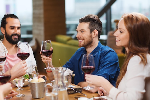 wypoczynek, jedzenie i napoje, świętowanie ludzi i koncepcja świąt - uśmiechnięci przyjaciele jedzący obiad i pijący czerwone wino w restauracji