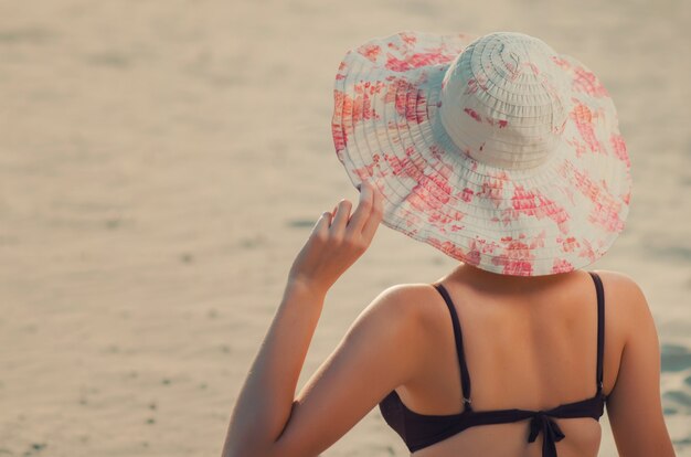 Wysoka szczupła kaukaska dziewczyna trzyma rękę przez krawędź czapki na plaży, siedzi na złotym piasku. Wypoczynek i relaks nad brzegiem morza (ocean, rzeka, jezioro) w letni i słoneczny dzień.