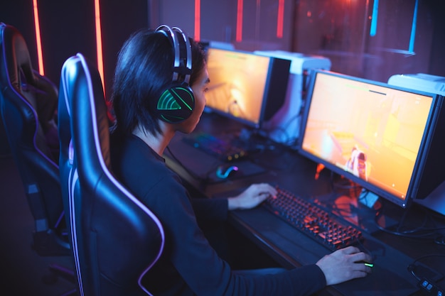 Wysoki kąt portret młodego mężczyzny azjatyckich, grając w gry wideo i nosząc słuchawki w ciemnym wnętrzu cyber sportu, kopia przestrzeń