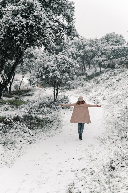 Wysoki kąt widok z tyłu anonimowej kobiety idącej z wyciągniętymi ramionami wzdłuż zaśnieżonej ścieżki i cieszącej się spacerami po lesie w zimie
