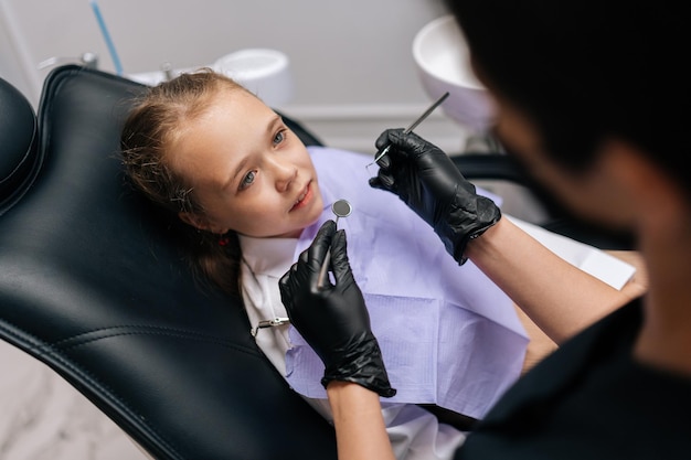 Wysoki kąt widoku przerażonej uroczej dziewczyny siedzącej na krześle dentystycznym i patrzącej ze strachem na nieznaną dentystkę trzymającą w rękach instrumenty dentystyczne