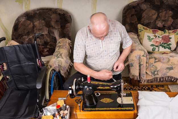 Wysoki kąt widzenia starszego mężczyzny pracującego w staromodnej maszynie do szycia w salonie w domu z wózkiem inwalidzkim w pobliżu