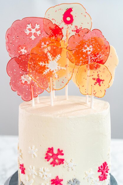 Wysoki okrągły tort z włoskim lukrem buttercream ozdobiony płatkami śniegu z kremówki i zwieńczony dużymi różowo-białymi lizakami na szarym tle.