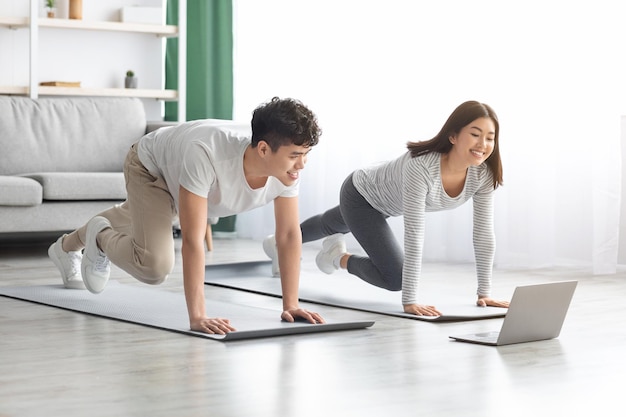 Wysportowana azjatycka rodzina ćwicząca razem w domu, rozciągająca się na macie fitness, patrząca na ekran laptopa, mająca zajęcia online, wnętrze salonu, miejsce do kopiowania. Sportowa czarna para trenuje razem