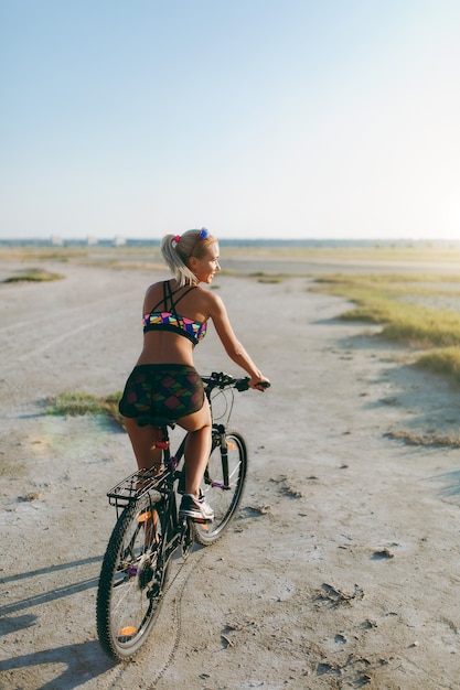 Wysportowana Blondynka W Kolorowym Garniturze Jeździ Na Rowerze Po Pustyni W Słoneczny Letni Dzień. Koncepcja Fitness. Widok Z Tyłu