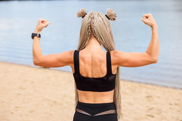 Wysportowana kobieta z fryzurą afro warkoczy pokazuje mięśnie ramion stojące plecami na zewnątrz na plaży latem
