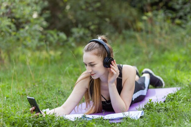 Wysportowana szczupła kobieta leży na macie słucha muzyki w słuchawkach i używa smartfona w parku Zdrowy styl życia
