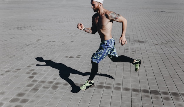 Wysportowany mężczyzna z nagim torsem z tatuażami i opaską na głowie, ubrany w czarne legginsy i niebieskie szorty biega po płytach chodnikowych w ciepły słoneczny dzień.