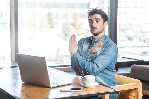 Wystarczająco! Widok Z Boku Portret Ostrzegawczy Agresywny Brodaty Młody Freelancer W Niebieskiej Koszuli Dżinsowej Siedzi W Kawiarni I Nawiązuje Wideorozmowę Na Laptopie Ze Skrzyżowanymi Podniesionymi Rękami Jak Gest Stop. Wnętrz,