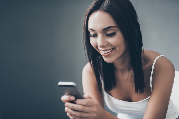 Wysyłam SMS-y do przyjaciela. Atrakcyjna młoda kobieta trzymająca inteligentny telefon i patrząca na niego z uśmiechem, siedząc przy szarej ścianie