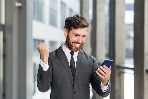 Wyszedł biznesmen świętuje osiągnięcie, patrząc na smartfona na świeżym powietrzu. Udany biznes człowiek świętuje wygraną z telefonu komórkowego na zewnątrz. Wesołych dobrych wieści. Udana transakcja, zakład początkowy, handel