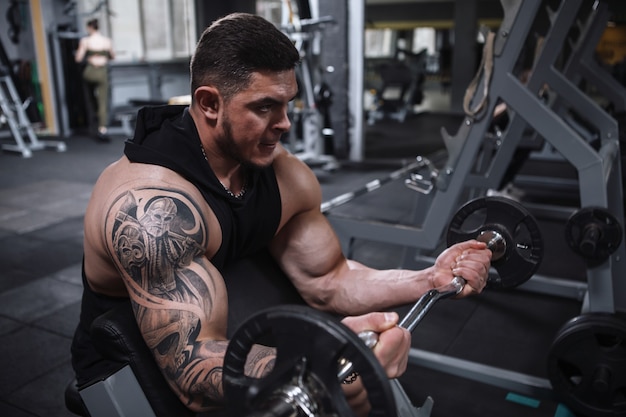 Wytatuowany wytatuowany sportowiec robi uginanie bicepsów ze sztangą na siłowni