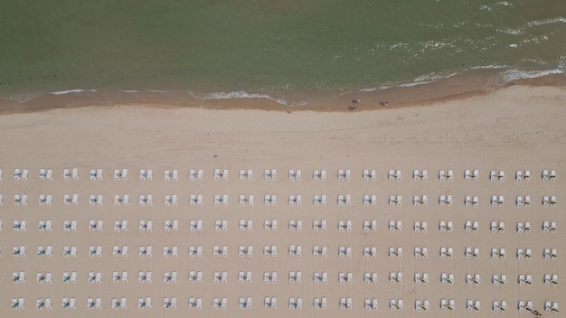 Wzdłuż wybrzeża ciągnie się piaszczysta plaża ozdobiona pustymi leżakami. Widok z lotu ptaka