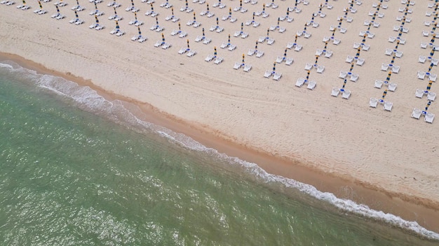 Wzdłuż wybrzeża ciągnie się piaszczysta plaża ozdobiona pustymi leżakami. Widok z lotu ptaka