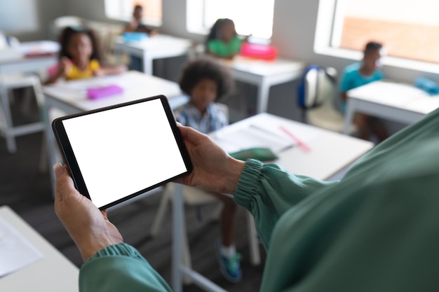 Z bliska młoda kaukazyjska nauczycielka używająca cyfrowego tabletu z wielonarodowymi uczniami w tle