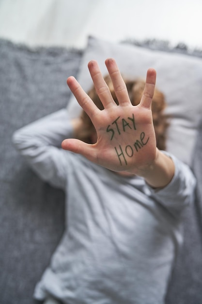 Z góry anonimowe dziecko pokazujące rękę z napisem „Zostań w domu”, leżące na kocu i poduszce podczas pandemii w domu