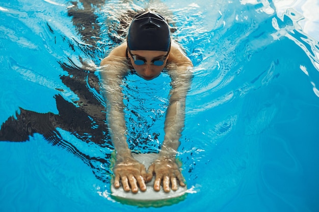 Z góry pływaczka w czapce i okularach pływająca z piankową deską w basenie podczas treningu