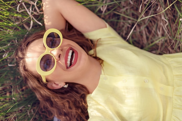 Z góry stylowa roześmiana kobieta w żółtych okularach przeciwsłonecznych i sukience leżąca na zielonej łące i ciesząca się słońcem w letni dzień