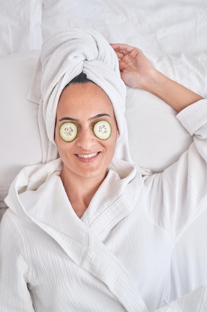 Z góry zrelaksowana kobieta w szlafroku i ręczniku z plasterkami ogórka na oczach, spoczywająca na łóżku podczas zabiegu pielęgnacji skóry