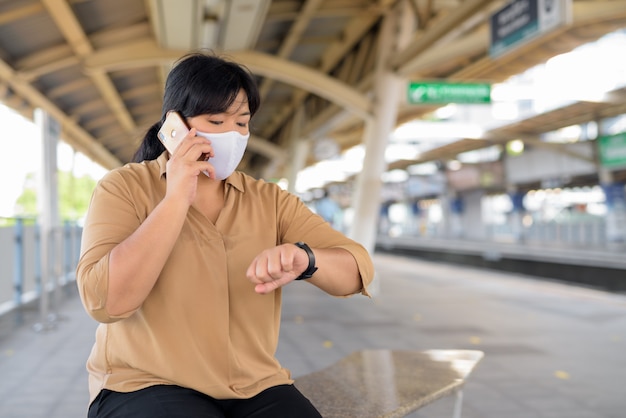 Z nadwagą Azjatycka kobieta rozmawia przez telefon i sprawdza czas przy niebo pociągiem z maską