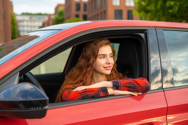 Z promieniującym uśmiechem młoda kobieta z rudymi włosami siedzi w czerwonym samochodzie patrząc przez otwarty