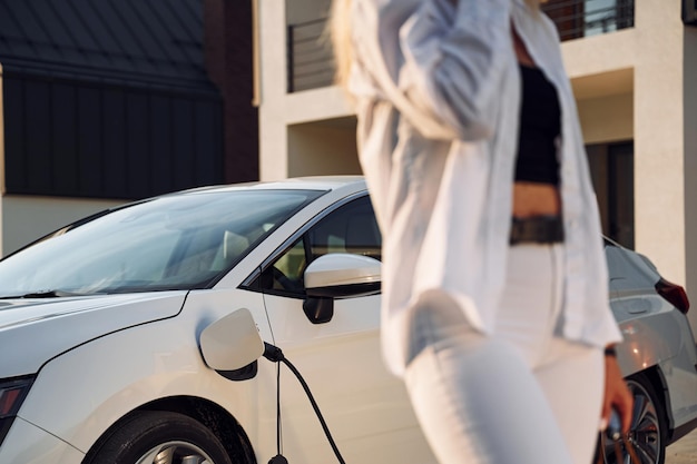 Z torbą sopping Młoda kobieta w białym ubraniu jest ze swoim samochodem elektrycznym w ciągu dnia
