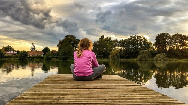 Zdjęcie z tyłu dziewczyna siedząca na molo nad jeziorem na tle nieba