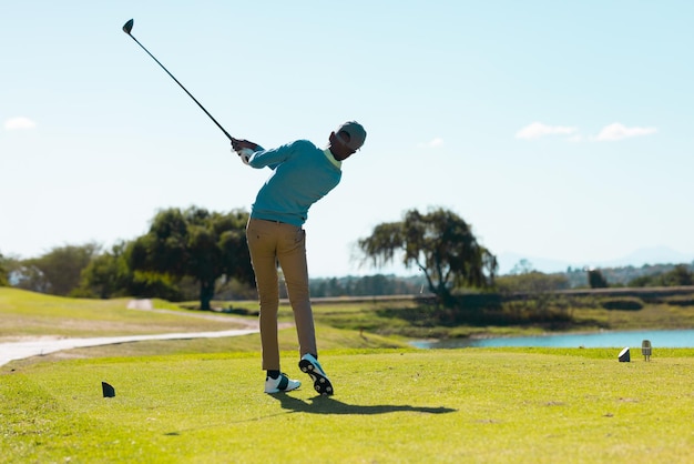 Zdjęcie z tyłu widok afroamerykańskiego młodego mężczyzny strzelającego do golfa na trawiastym terenie na polu golfowym przeciwko niebu