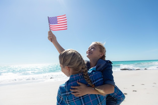 Z tyłu widok białej dziewczyny podnoszącej amerykańską flagę z matką, stojącej na plaży z niebieskim niebem i morzem w tle, obejmującej się i uśmiechającej się