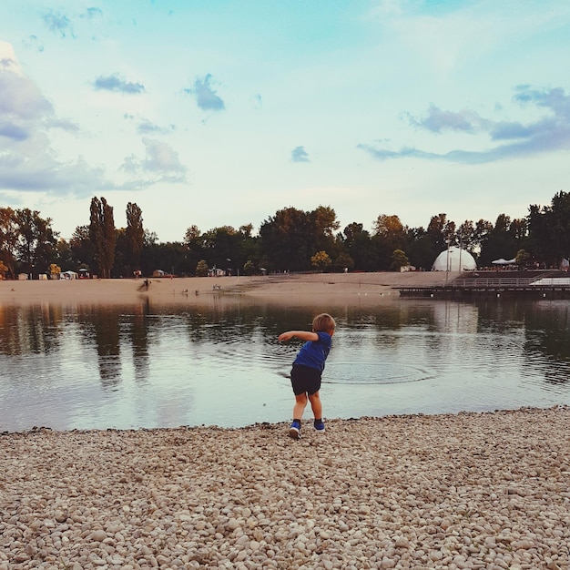 Zdjęcie z tyłu widok chłopca rzucającego kamienie w jezioro na tle nieba