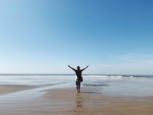 Z tyłu widok kobiety z podniesionymi ramionami stojącej na plaży na tle niebieskiego nieba
