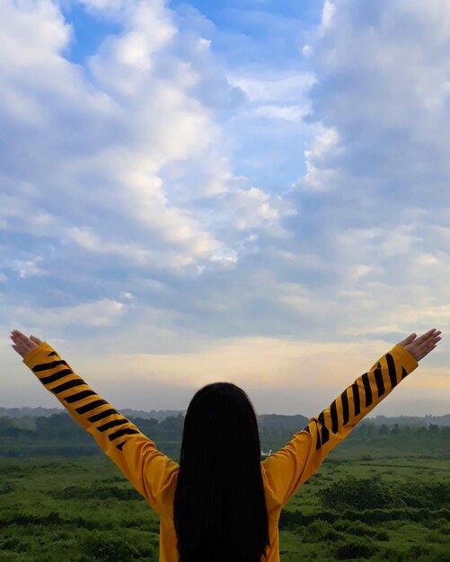 Zdjęcie z tyłu widok kobiety z podniesionymi ramionami stojącej na polu na tle chmurnego nieba