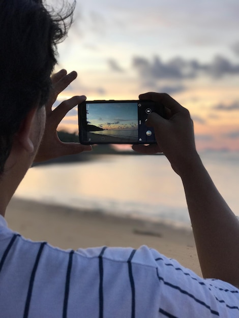 Z tyłu widok mężczyzny fotografującego przez smartfon na plaży podczas zachodu słońca