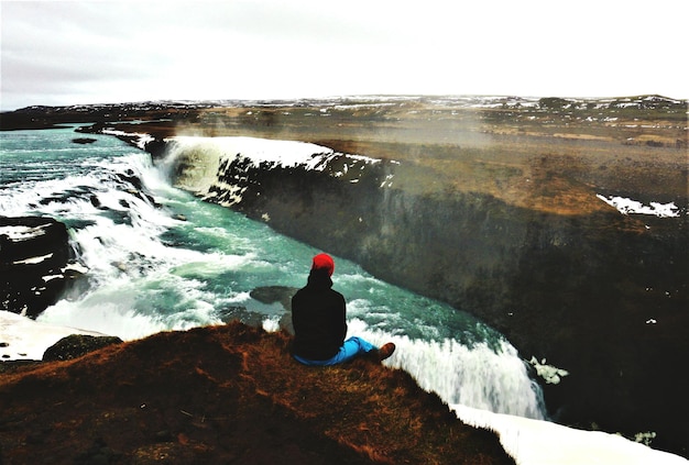 Zdjęcie z tyłu widok osoby siedzącej na skale obok rzeki na czystym niebie